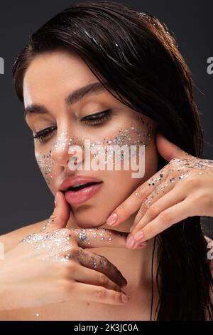giovane donna brunetta con scintillante glitter su guance e mani che toccano il viso isolato su grigio, immagine stock Foto Stock