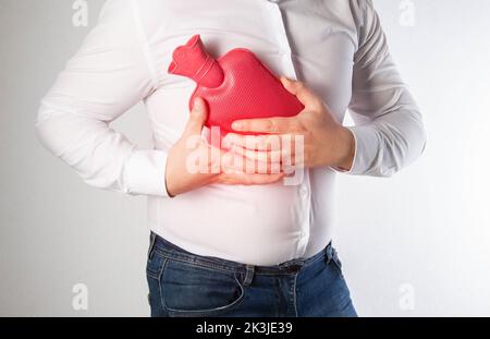 Un uomo in una camicia bianca tiene un cuscinetto di riscaldamento rosso con acqua calda vicino al petto delle costole. Trattamento della nevralgia intercostale e del nervo pizzicato Foto Stock
