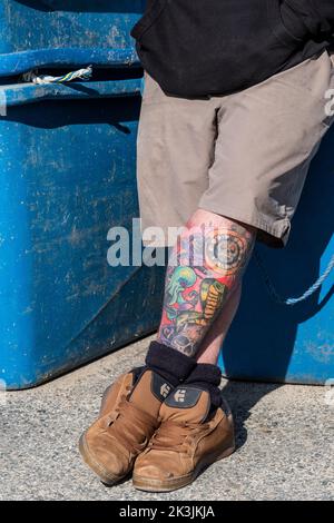 Un uomo che si rilassa contro i bidoni di stoccaggio con le gambe tatuate incrociate. Foto Stock