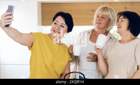 Felice donna di mezza età con smartphone che prende selfie a casa e bere tè o caffè. Gruppo di donne mature felici che prendono auto-ritratto. Foto Stock