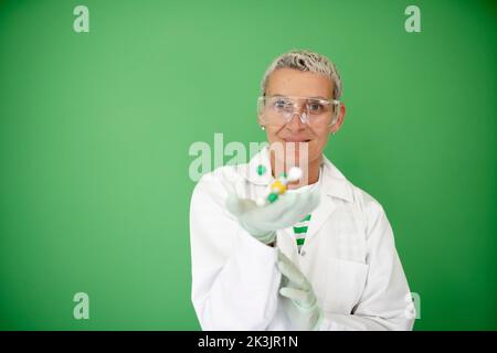 scienziato femminile con mantello bianco tiene in mano il modello di molecola e si trova di fronte allo sfondo verde Foto Stock