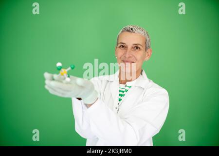 scienziato femminile con mantello bianco tiene in mano il modello di molecola e si trova di fronte allo sfondo verde Foto Stock