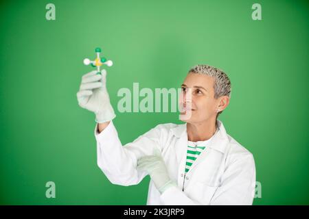 scienziato femminile con mantello bianco tiene in mano il modello di molecola e si trova di fronte allo sfondo verde Foto Stock