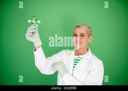 scienziato femminile con mantello bianco tiene in mano il modello di molecola e si trova di fronte allo sfondo verde Foto Stock