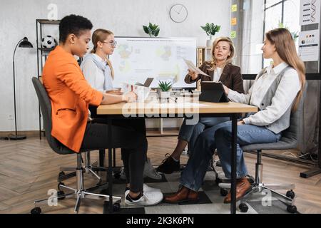 Donne d'affari concentrate nell'abbigliamento formale che trascorrono il tempo in ufficio per monitorare la situazione del mercato finanziario. Economisti donne qualificati seduti alla scrivania con un moderno notebook, un tablet e un enorme display. Foto Stock
