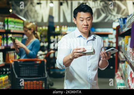 Un giovane asiatico è in piedi in un supermercato, scegliendo i pattini per la sua moglie, ragazza. Non sa quale scegliere, si alza pensieroso, confuso, tenendo il telefono in mano. Foto Stock