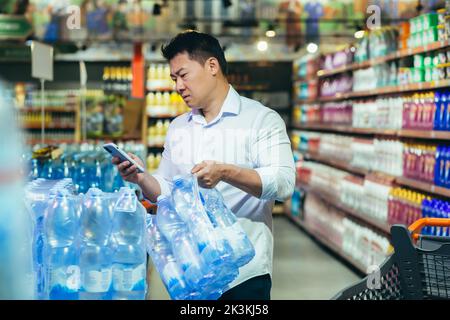 Acquisto all'ingrosso. Un giovane asiatico serio compra un pallet di acqua minerale in un supermercato. Tiene il telefono in mano, guarda il telefono, utilizza il self-service. Foto Stock