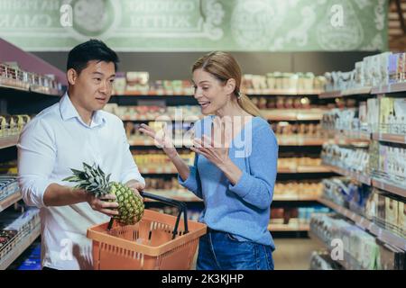 Conflitto di famiglia nel negozio. Giovane coppia interracial che sostiene in un supermercato. Una donna grida ad un uomo asiatico che ha speso i soldi in frutta costosa. Un uomo è confuso con un cesto nelle sue mani. Foto Stock