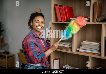 Felice giovane donna nera con spazzola pulisce polvere sui mobili, ama la pulizia nel soggiorno interno Foto Stock