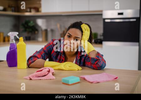 Ragazza afro-americana stanca infelice in guanti di gomma a tavola con i rifornimenti di pulizia Foto Stock