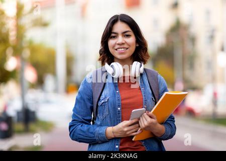 Studente moderno. Sorridente giovane ragazza araba con libri degli esercizi e smartphone in posa all'aperto Foto Stock