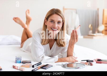 Donna felice matura in accappatoio sdraiata sul letto e tenendo il rossetto, guardando la macchina fotografica e sorridendo all'interno della camera da letto Foto Stock