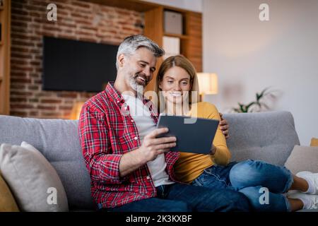 Allegra coppia matura europea hanno videochiamate in remoto, chattando dal tablet seduti sul divano Foto Stock