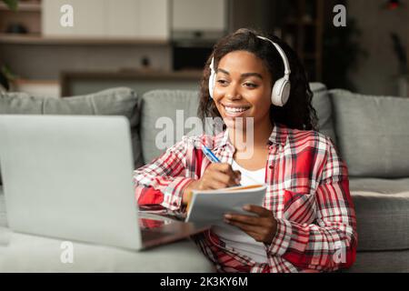 Allegra giovane afroamericana in cuffie wireless prendere appunti, guardare video lezione sul computer portatile Foto Stock