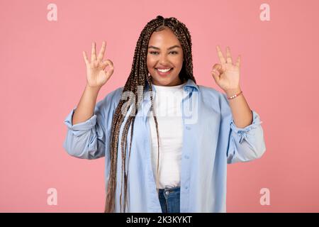 Allegro Nero Femminile gesturing OK con entrambe le mani, sfondo rosa Foto Stock