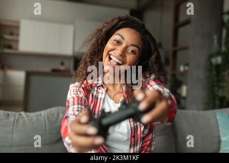 Felice eccitato giovane afroamericana donna con bocca aperta giocare gioco online con joystick, seduto sul divano Foto Stock