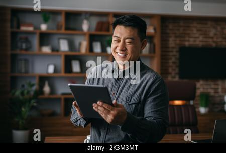 Sorridente uomo coreano maturo che digita sul tablet, parla con il cliente, analizza i dati di avvio all'interno dell'ufficio Foto Stock