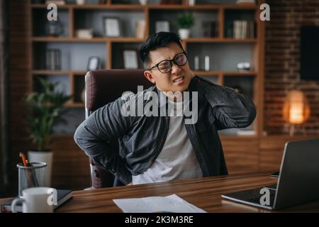 Uomo giapponese adulto stanco in occhiali che soffrono di una seduta lunga in una postura sbagliata da parte della schiena e del collo ferito Foto Stock