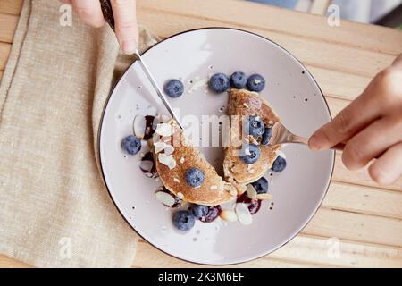 Donna con posate mangiare frittelle tradizionali con mirtilli freschi e sciroppo di agave su un piatto. Colazione all'aperto sulla terrazza. Foto Stock