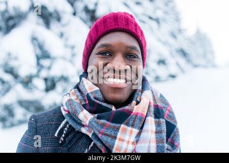 uomo afro-americano in cappello rosso ed elegante cappotto a plaid guardare la macchina fotografica con il sorriso bianco-neve al coperto nel parco Foto Stock