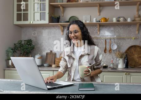 Giovane bella donna ispanica con capelli ricci che lavora a casa utilizzando un computer portatile, donna in cucina sorridente e felice freelancer remoto lavoro. Foto Stock