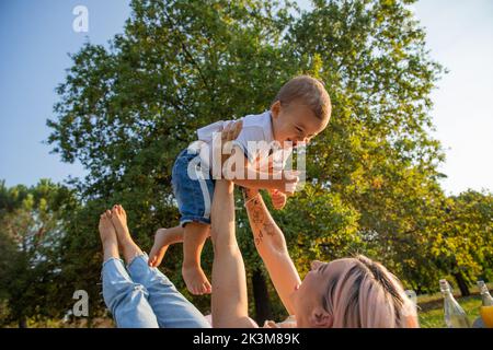 La giovane madre gioca con il figlio sollevandolo nell'aria durante il picnic. Giorno della mamma. Foto Stock