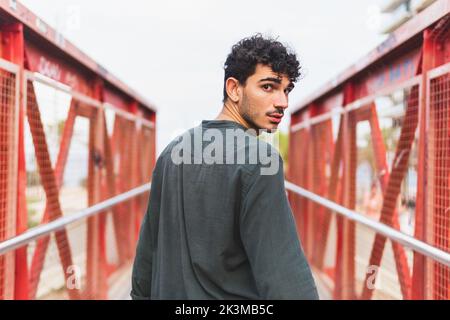 Vista posteriore di un giovane maschio ispanico in camicia nera che cammina sul ponte e guarda la macchina fotografica sopra la spalla mentre cammina sul ponte pedonale sulla spiaggia Foto Stock