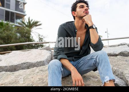 Turista maschile ispanico in abiti casual appoggiandosi a mano e pensando mentre si siede sulle rocce il giorno del fine settimana in spiaggia Foto Stock