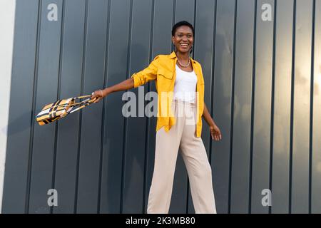 Allegro sicuro adulto afroamericano femmina con i capelli scuri corti in vestito elegante sorridente e guardando via mentre levandosi in piedi contro la parete grigia con Foto Stock