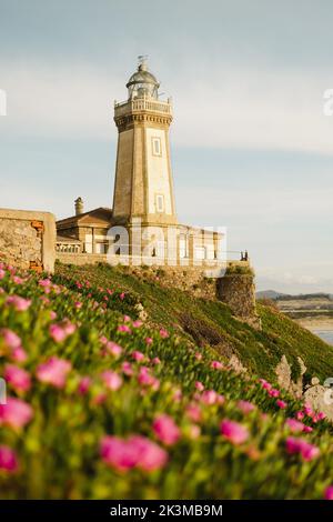 Aged San Juan Faro situato su collina erbosa vicino a fiori rosa e il mare contro cielo nuvoloso in Aviles, Spagna Foto Stock