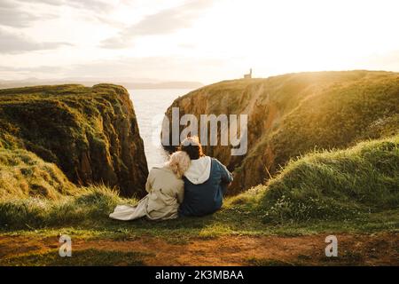 Vista posteriore dell'uomo anonimo e della donna in outerwear appoggiati l'uno sull'altro e osservando il mare mentre si siede sulla scogliera al tramonto ad Aviles, Spagna Foto Stock