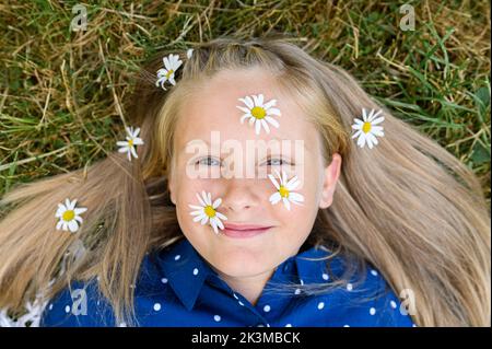 Vista dall'alto della ragazza bionda con fiori freschi di margherita sul viso e sui capelli guardando la macchina fotografica con il sorriso mentre sdraiato sull'erba il giorno d'estate Foto Stock