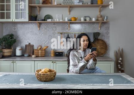 Seria e premurosa donna latino-americana in cucina utilizza il telefono, donna legge con attenzione un messaggio da un'applicazione online Foto Stock
