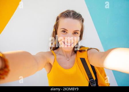 Giovane donna allegra con zaino che guarda la fotocamera mentre prende selfie sulla moderna strada cittadina Foto Stock