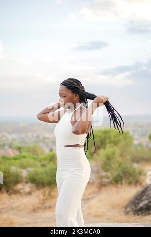 Donna afroamericana in abiti bianchi che toccano capelli lunghi intrecciati contro cielo nuvoloso il giorno d'estate in campagna Foto Stock