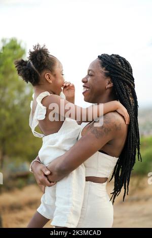 Vista laterale di felice madre afro-americana in abiti bianchi con capelli intrecciati sorridente e abbracciante figlia, mentre in piedi contro alberi lussureggianti e nuvoloso Foto Stock