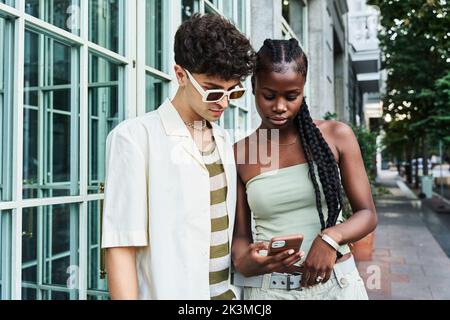 Giovane afroamericana con lunghe trecce che indossano la parte superiore e che condividono il telefono cellulare al bel ragazzo in occhiali da sole in piedi sulla strada Foto Stock