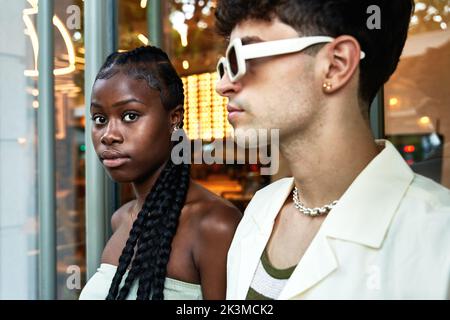Giovane donna nera con capelli intrecciati che guarda la macchina fotografica mentre si trova vicino a un elegante ragazzo contro una parete di vetro Foto Stock
