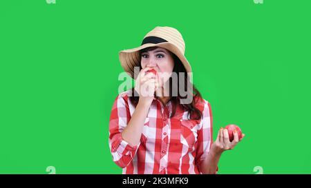 divertente, sorridente femmina contadina in camicia a scacchi e cappello, tenendo in mano due grandi mele acute, mangia, morde, rughe il viso, su sfondo verde in studio. Cibo sano al vostro tavolo. Foto di alta qualità Foto Stock