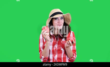 divertente, sorridente femmina contadina in camicia a scacchi e cappello, tenendo in mano due grandi mele acute, mangia, morde, rughe il viso, su sfondo verde in studio. Cibo sano al vostro tavolo. Foto di alta qualità Foto Stock