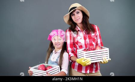 ritratto di famiglia di contadini, madre e figlia tenendo in mano scatole di legno con mele biologiche rosse mature, sorridente, su sfondo grigio in studio. Foto di alta qualità Foto Stock