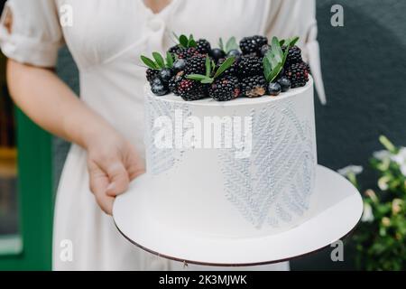 Un pasticciere in abito bianco tiene una torta bianca con bacche nelle mani su un piedistallo. Foto Stock