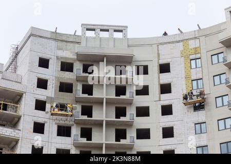 Processo di isolamento delle pareti esterne in un edificio a più piani in costruzione. I lavoratori in una culla arancione da costruzione attaccano le lana minerale Foto Stock