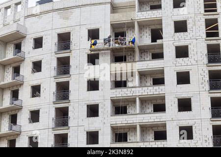 Processo di isolamento delle pareti esterne in un edificio a più piani in costruzione. I lavoratori in una culla arancione da costruzione attaccano le lana minerale Foto Stock