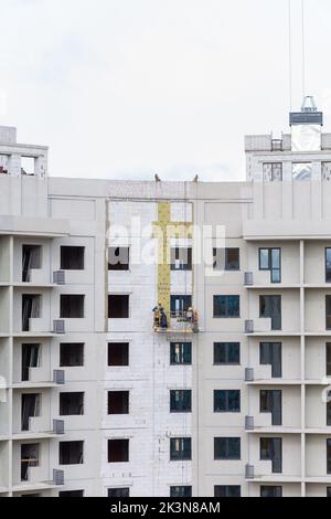 Processo di isolamento delle pareti esterne in un edificio a più piani in costruzione. I lavoratori in una culla arancione da costruzione attaccano le lana minerale Foto Stock