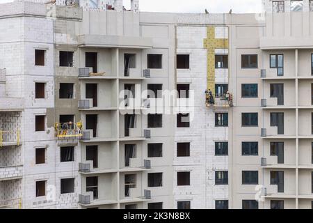 Processo di isolamento delle pareti esterne in un edificio a più piani in costruzione. I lavoratori in una culla arancione da costruzione attaccano le lana minerale Foto Stock