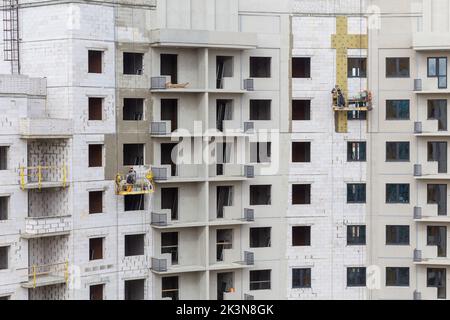 Processo di isolamento delle pareti esterne in un edificio a più piani in costruzione. I lavoratori in una culla arancione da costruzione attaccano le lana minerale Foto Stock