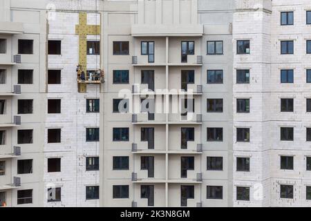 Processo di isolamento delle pareti esterne in un edificio a più piani in costruzione. I lavoratori in una culla arancione da costruzione attaccano le lana minerale Foto Stock