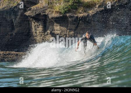 Un giovane surfista maschile naviga su una grande onda vuota di fronte alle scogliere di Piha Beach, Auckland, Nuova Zelanda. Foto Stock