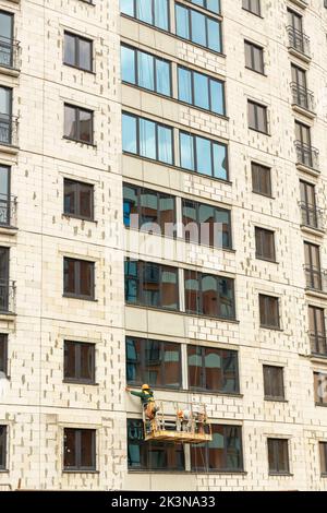 Processo di isolamento delle pareti esterne in un edificio a più piani in costruzione. I lavoratori in una culla arancione da costruzione attaccano le lana minerale Foto Stock